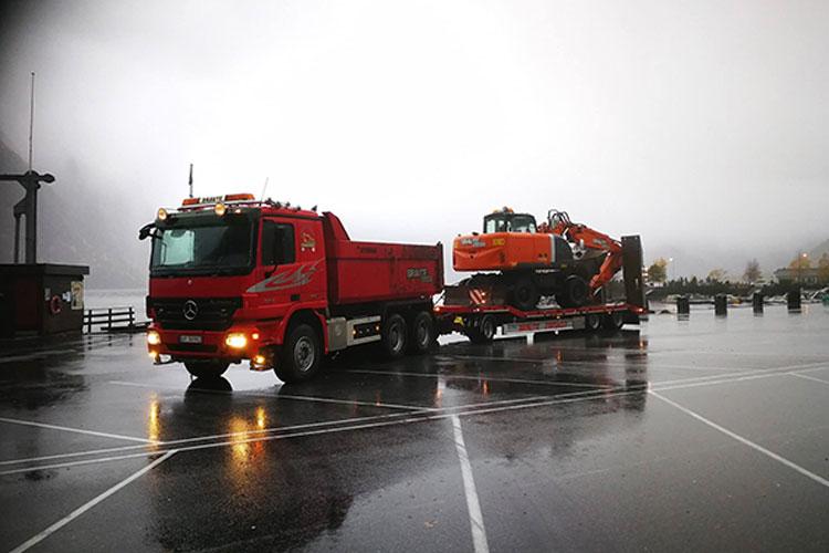 Rød lastebil med gravemaskin på tilhenger, parkert på en våt plass i regnvær.