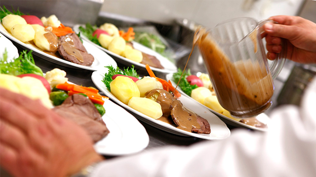En kokk heller saus over tallerkener med kjøtt, poteter og grønnsaker i et restaurantkjøkken.