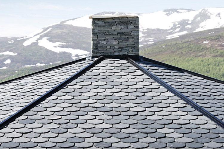 Tak med skiferstein og mursteinpipe, med snødekte fjell i bakgrunnen.