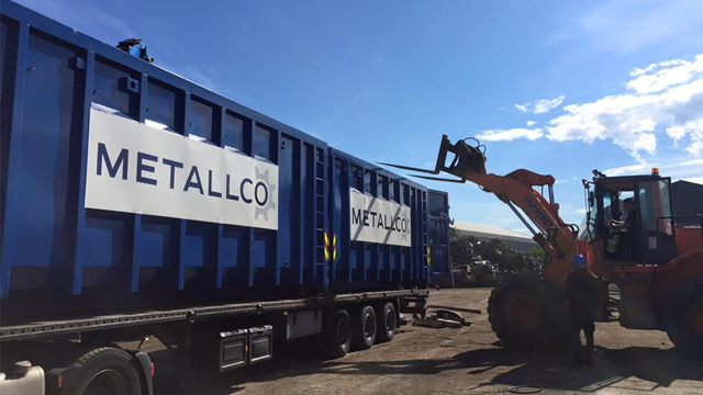 Blå containere med Metallco-logo på en lastebil, med en gravemaskin som laster materialer.