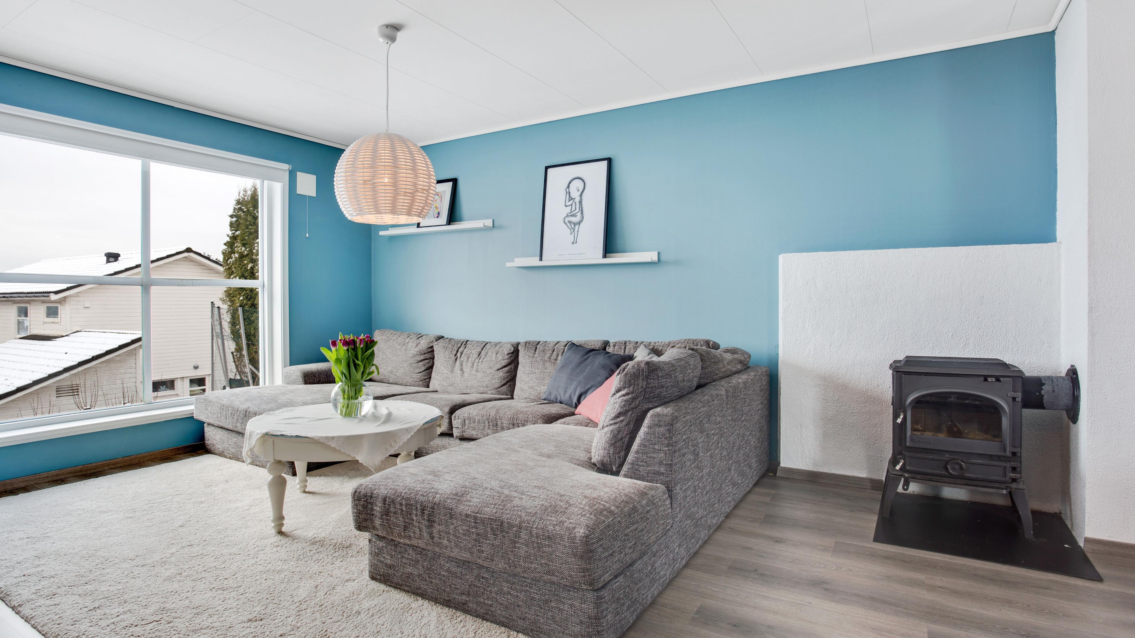 Lys stue med blå vegger, grå sofa, bord, og peis, samt store vinduer som slipper inn naturlig lys.