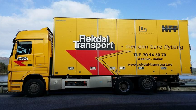 Gul lastebil fra Rekdal Transport med teksten "men enn bare flytting" på siden, parkert ved veien.