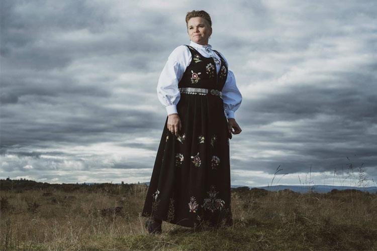 Kvinne iført bunad står på en åpen slette under en dramatisk skyhimmel.