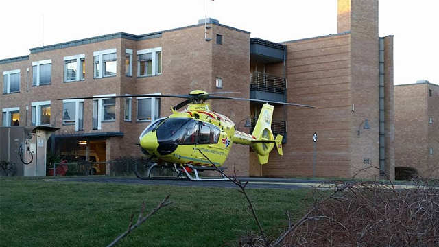 En gul ambulansehelikopter står parkert foran en murbygning med flere vinduer.