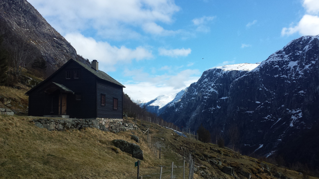 Et svart hus i fjellandskap med snødekte topper og blå himmel i bakgrunnen.