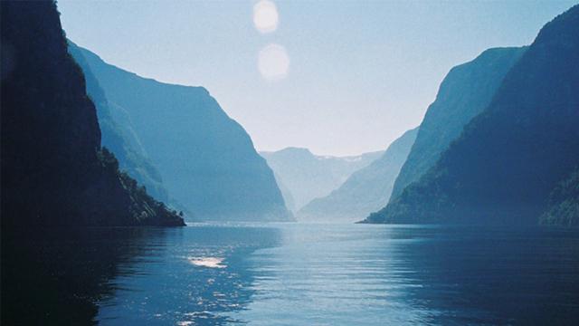 En vakker fjord med høye fjell og stille vann under en klar blå himmel. Sollys reflekteres på vannoverflaten.