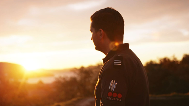 En mann i uniform ser mot solnedgangen, med et vakkert landskap i bakgrunnen.
