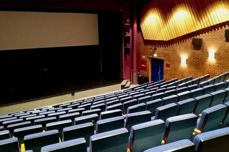 En tom kinosal med blå seter og en stor skjerm på scenen, belyst av varme vegglamper.