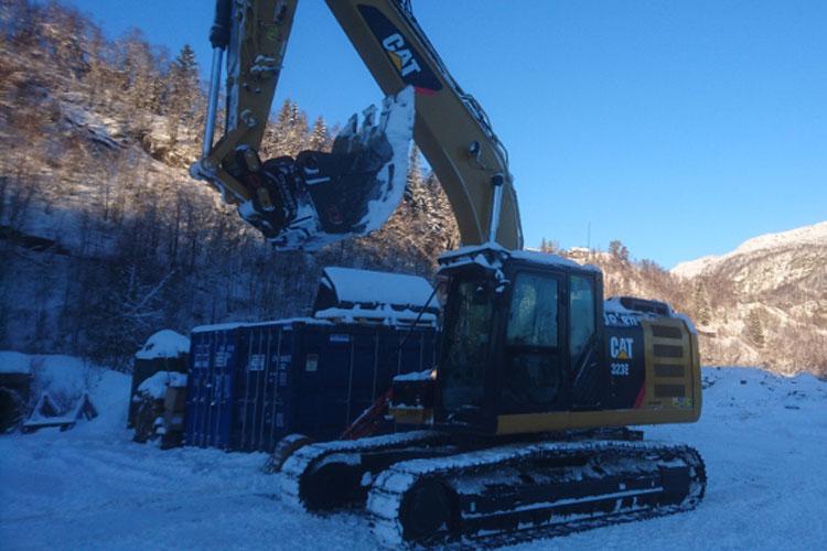 Gravemaskin fra CAT i snødekt landskap, med containere i bakgrunnen og fjell i horisonten.