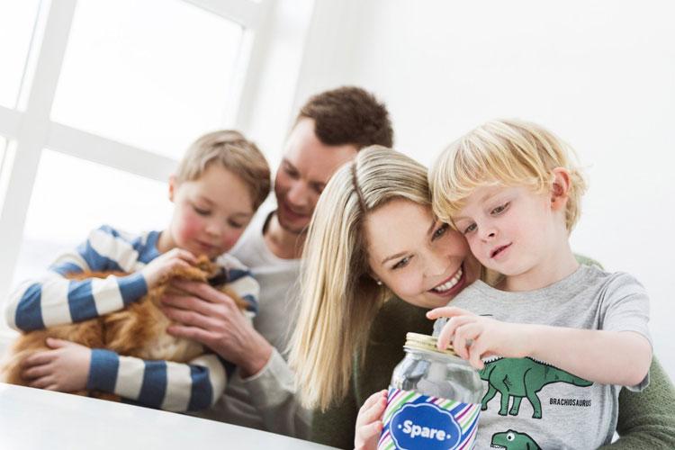 Familie med to barn som ser på sparebøsse, en katt i bakgrunnen, i et lyst rom med vinduer.