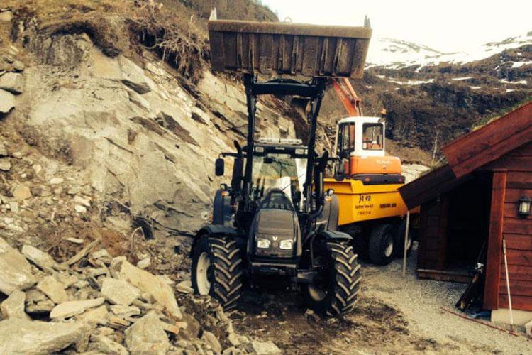 En traktor og en gravemaskin jobber på en steinete skråning ved en hytte i fjellet.