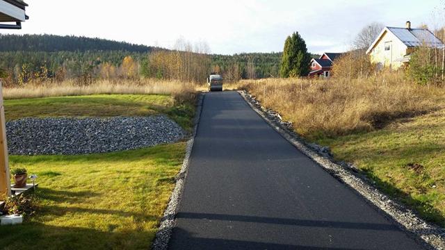 Ny asfaltlagt vei som fører gjennom et grønt landskap med trær og hus i bakgrunnen.