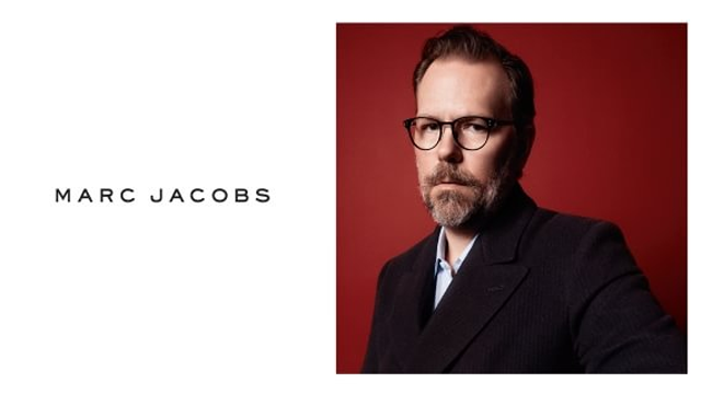 Portrett av Marc Jacobs med briller, iført en mørk dress mot en rød bakgrunn.