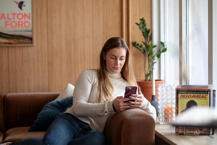 Kvinne sitter på en sofa og ser på telefonen sin, med planter og bøker i bakgrunnen.