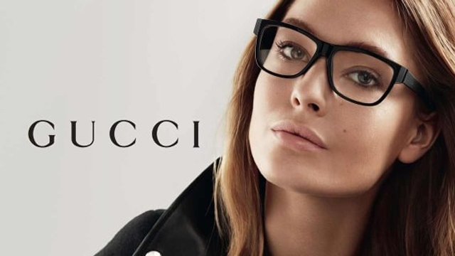 Modell med briller poserer foran en Gucci-logo, med et stilrent og moderne utseende.