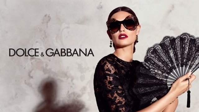 Kvinne med solbriller og svart vifte, iført en elegant svart kjole, med Dolce & Gabbana-logo i bakgrunnen.
