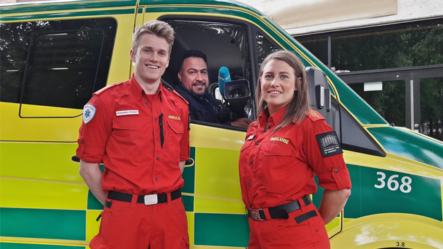 To personer i røde ambulanseuniformer smiler foran en ambulanse, med en sjåfør i førersetet.