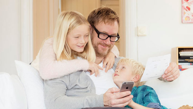 Far sitter på sofaen med to barn, en jente og en gutt, mens de ser på telefonen hans. De smiler og koser seg sammen.