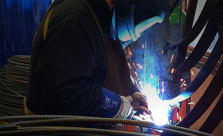 En sveiser i arbeid med lysbue, iført verneutstyr og hansker, omringet av sveiseutstyr og metalltråder.