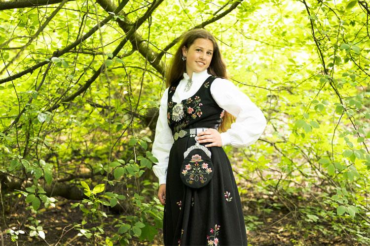Kvinne i tradisjonell bunad med broderier, står under grønne trær i en skog.