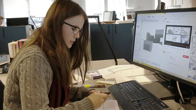 En kvinne med briller sitter ved et skrivebord og jobber med en datamaskin, omringet av papirer og kontorutstyr.