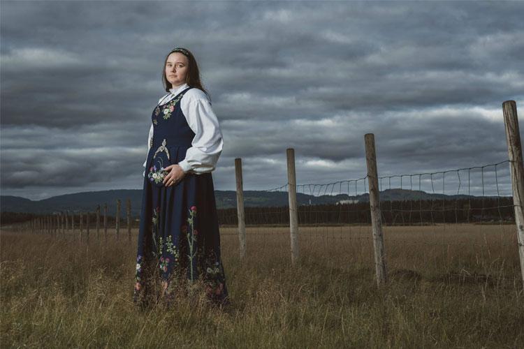 Kvinne i tradisjonell bunad står i et åpent landskap med gjerde og mørke skyer i bakgrunnen.