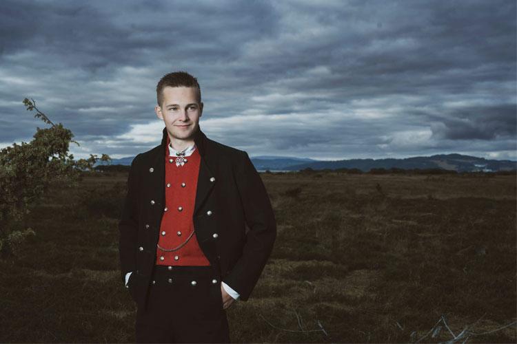 En mann i tradisjonell norsk bunad står ute i naturen med mørke skyer i bakgrunnen.