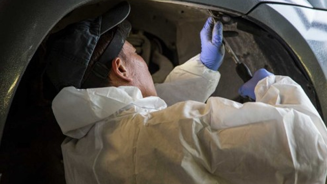 En person i hvit arbeidsdrakt og hansker jobber med bilreparasjon under bilen.