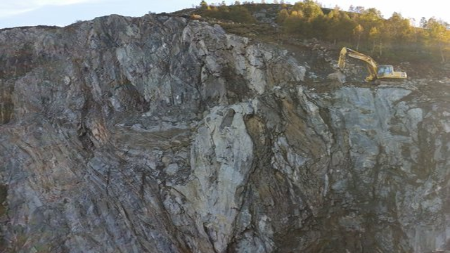 Grå fjellvegg med en gravemaskin som arbeider på toppen av skråningen i et steinbrudd.