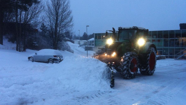 Traktor som ploger snø i en parkeringsplass med snødekte biler og bygninger i bakgrunnen.