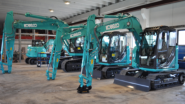 Tre Kobelco gravemaskiner i en lagerbygning, med tydelig grønn farge og moderne design.