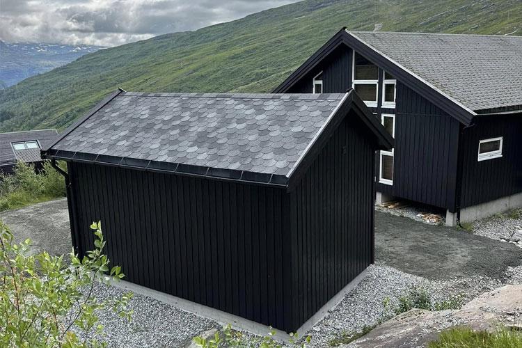 Moderne svart hytte med skråtak, beliggende i fjellandskap med grønn bakgrunn.