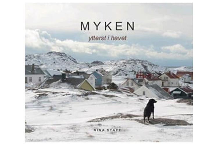 En svart hund står på en steinete bakgrunn med snødekte fjell og hus i Myken, ytterst i havet.