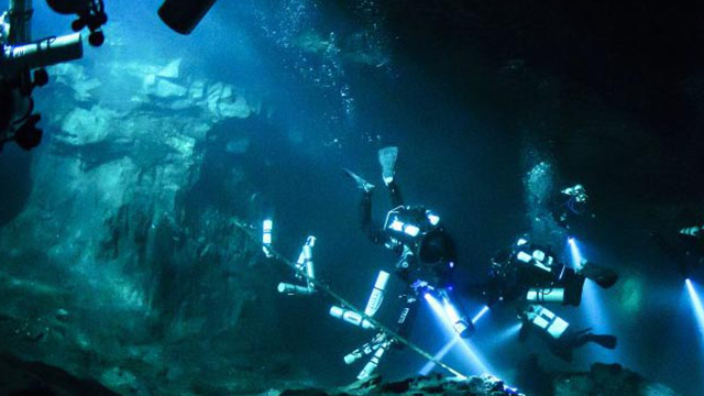 Dykkere utforsker en undervannsgrotte med lys i mørket, omringet av steinformasjoner og bobler.