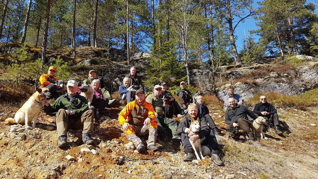 En gruppe mennesker og hunder sitter på en steinete bakke i skogen, omgitt av trær og sollys.