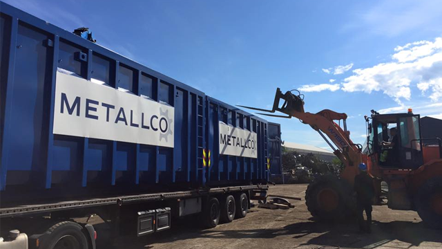 Blå containere med Metallco-logo lastes av en gaffeltruck på en byggeplass under klar himmel.