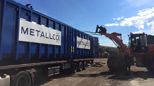 En gravemaskin laster av en blå container med Metallco-logo på en byggeplass under solfylt himmel.