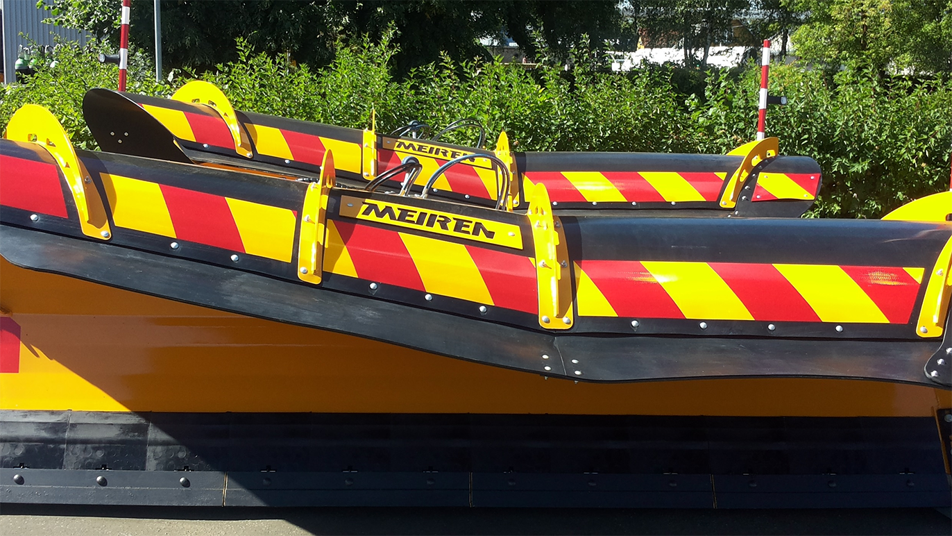 En stor snøplog med rød og gul stripe, parkert ved en grønn hekk.