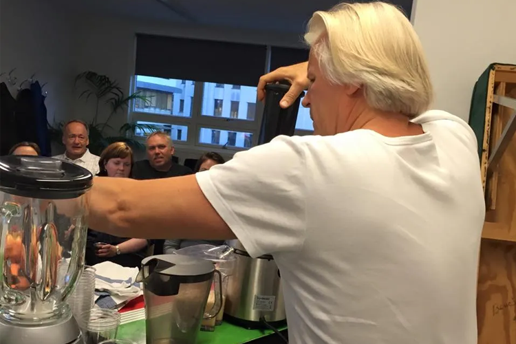 En person lager smoothie med en blender foran et publikum i et rom.