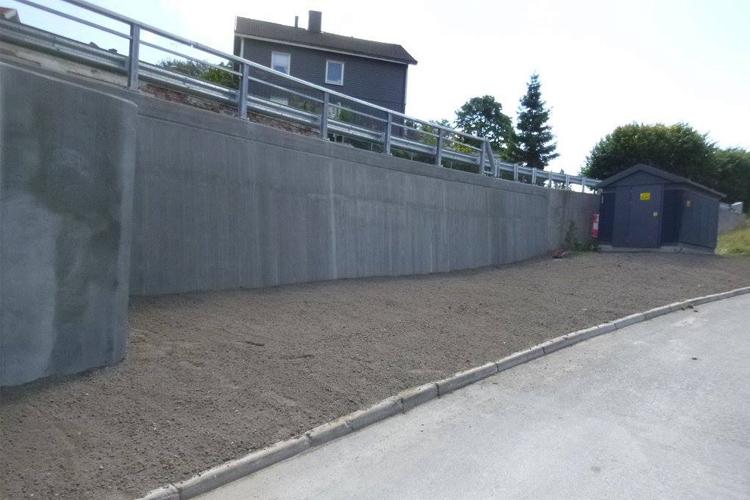 Betongvegg med rekkverk og en liten bygning ved siden av en gruslagt vei.