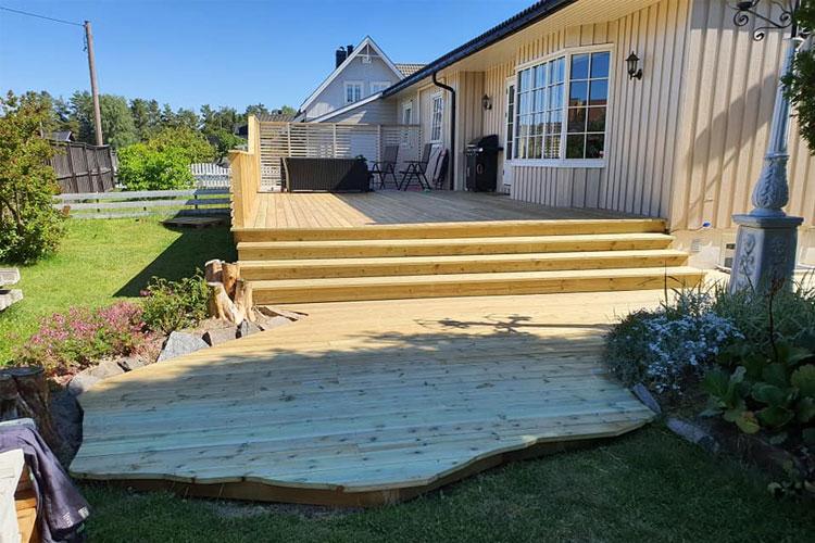 En ny treterrasse foran et hus med hage og blomster, med en hund som sitter på bakken.