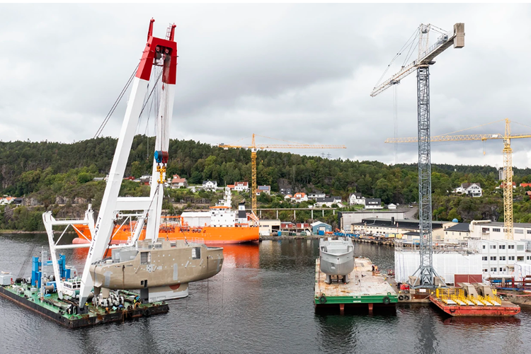 En stor kran heiser en båt i havnen, med flere byggekraner og skip i bakgrunnen.