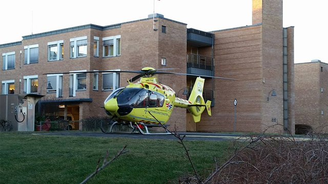 En gul ambulansehelikopter parkert foran en murbygning med grønt gress i forgrunnen.