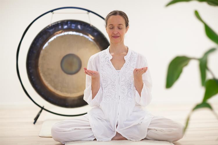 Kvinne i hvit klær med lukkede øyne sitter i meditasjon foran en gong. Grønne planter i bakgrunnen.
