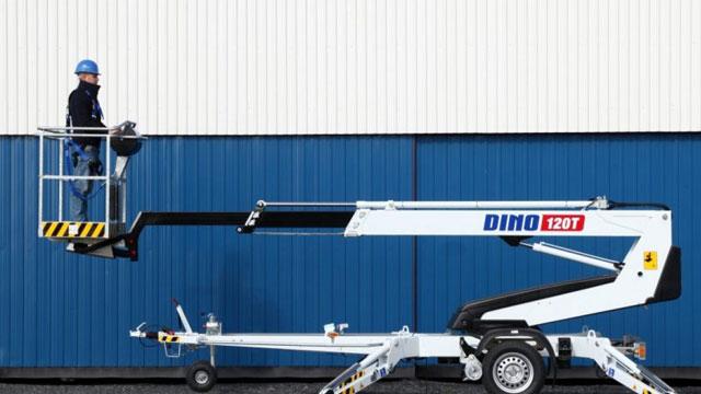 En person i hjelm står i en heisplattform på en DINO 120T arbeidsheis foran en blå vegg.