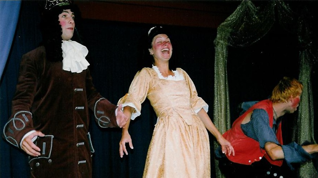 Tre skuespillere i kostymer på scenen, med en kvinne i en gul kjole og to menn i historiske drakter.
