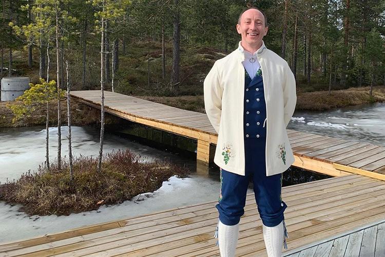 En mann i tradisjonell norsk bunad står på en trebro ved en innsjø med is og trær i bakgrunnen.