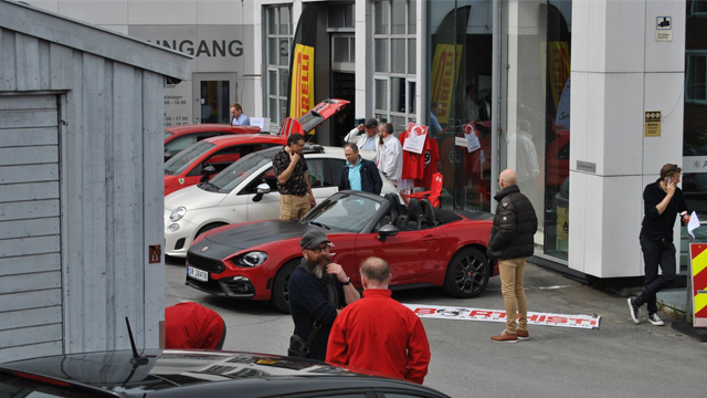 En rød sportsbil står parkert foran en bilforhandler, med folk som samles rundt og ser på biler.