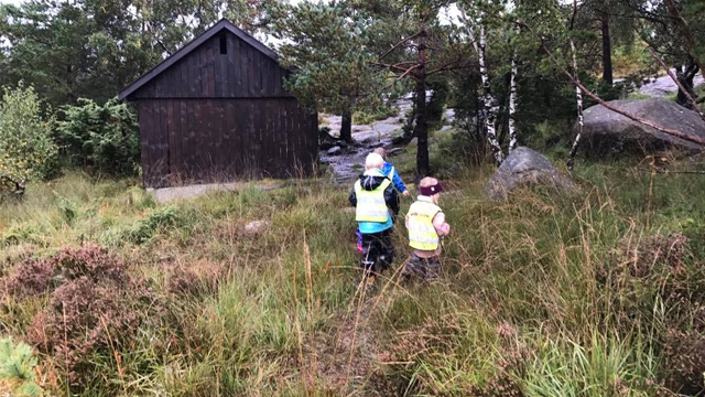 To barn i refleksvester går på en sti i skogen, med en hytte i bakgrunnen og steiner rundt.