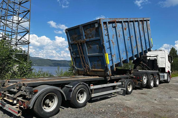 En lastebil med en blå tippcontainer står parkert ved en innsjø under en klar himmel.